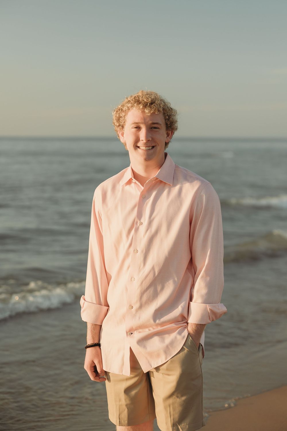 Smiling curly-haired young man in a light pink shirt and khaki shorts stands on the shoreline with gentle waves and a soft sky behind.