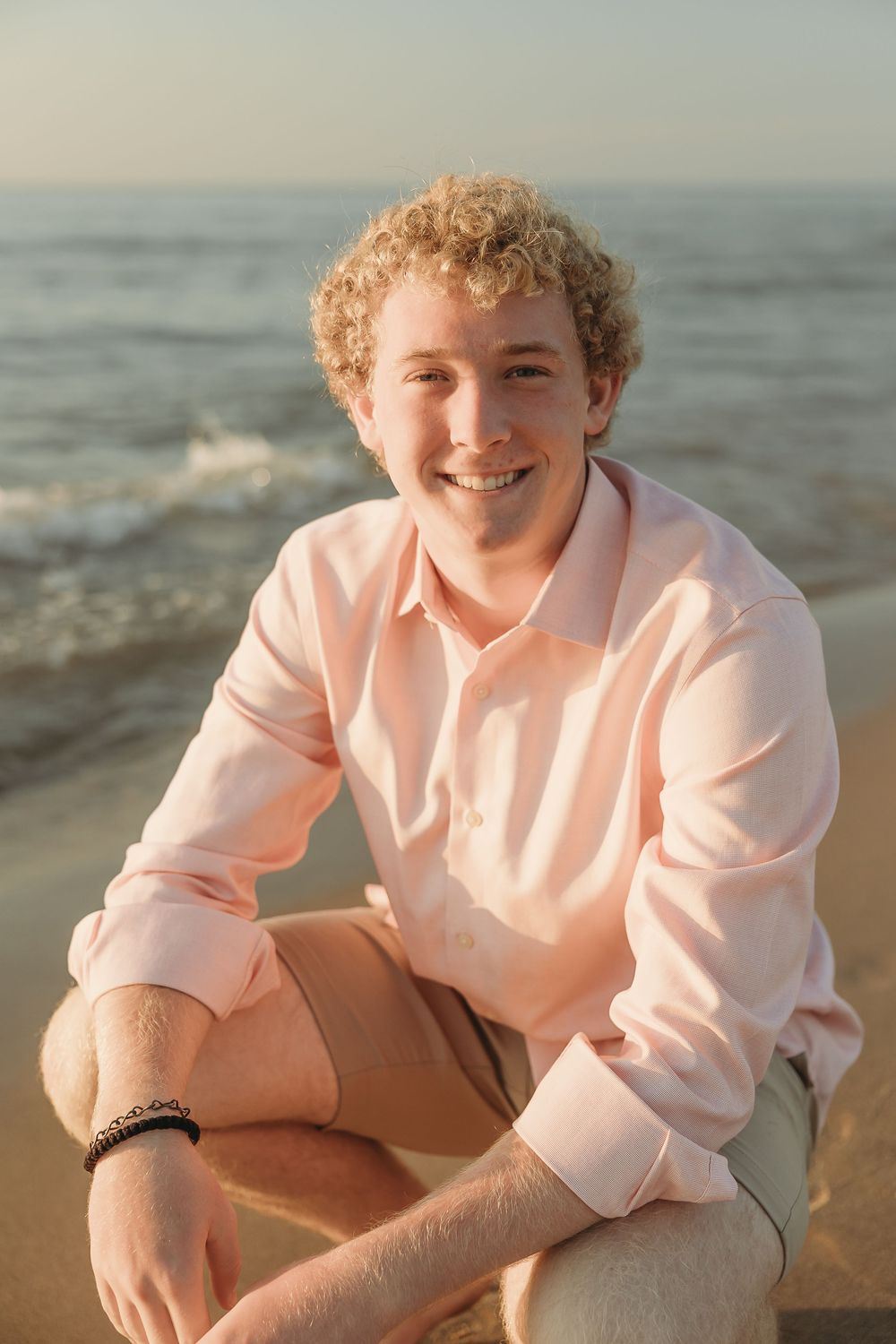 Smiling young man with curly blond hair in a pink button-up shirt and khaki shorts crouches on a sandy beach at sunset.