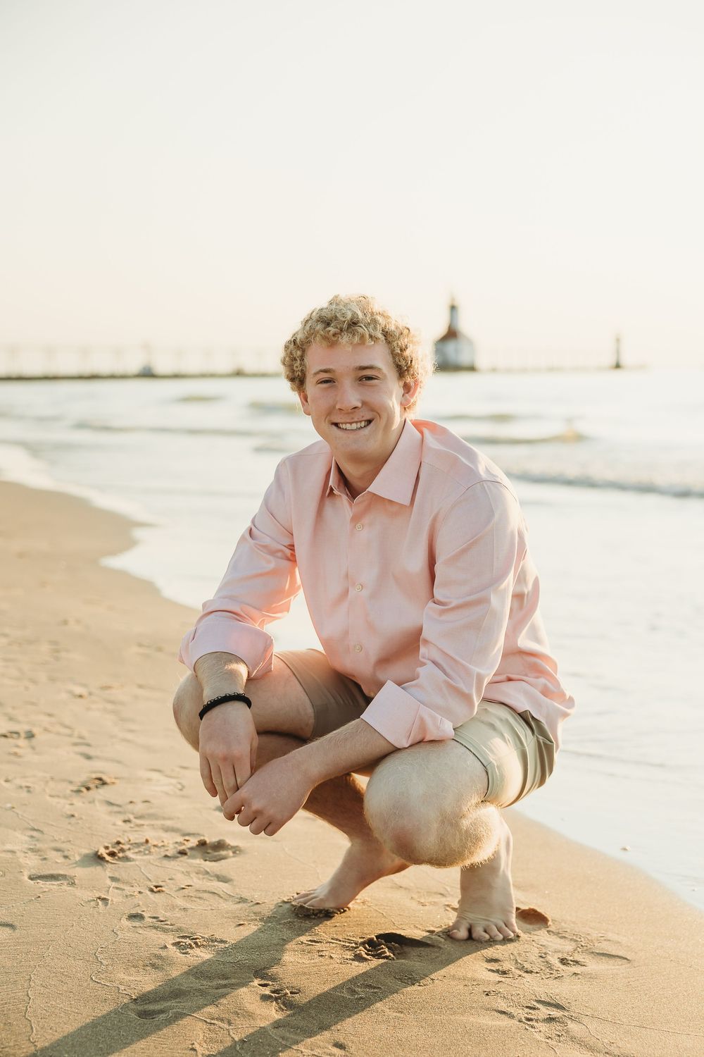 Smiling young man in a pink button-up shirt crouches on a sandy beach at sunset with a lighthouse and calm lake waves behind him.