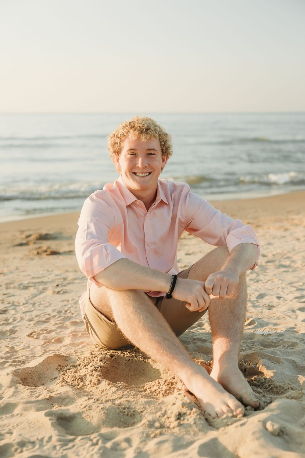 Smiling young man with curly blond hair in a pink shirt and khaki shorts sits barefoot on a sandy beach by the ocean at sunset.