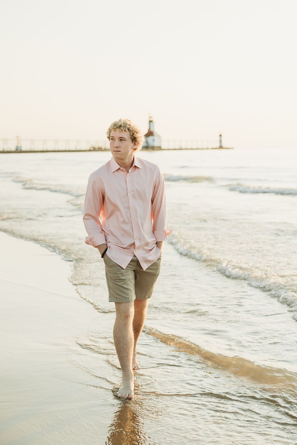 Young man with curly blonde hair walks barefoot through shallow waves along a sandy beach with a lighthouse pier in the distance.