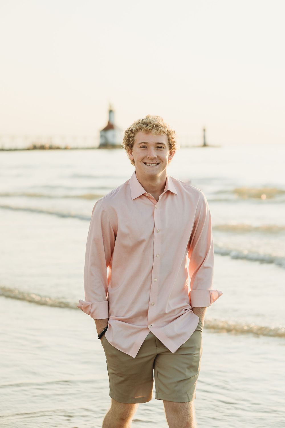 Smiling young man with curly blond hair in a pink button-up shirt and khaki shorts stands in shallow water with a lighthouse behind him.