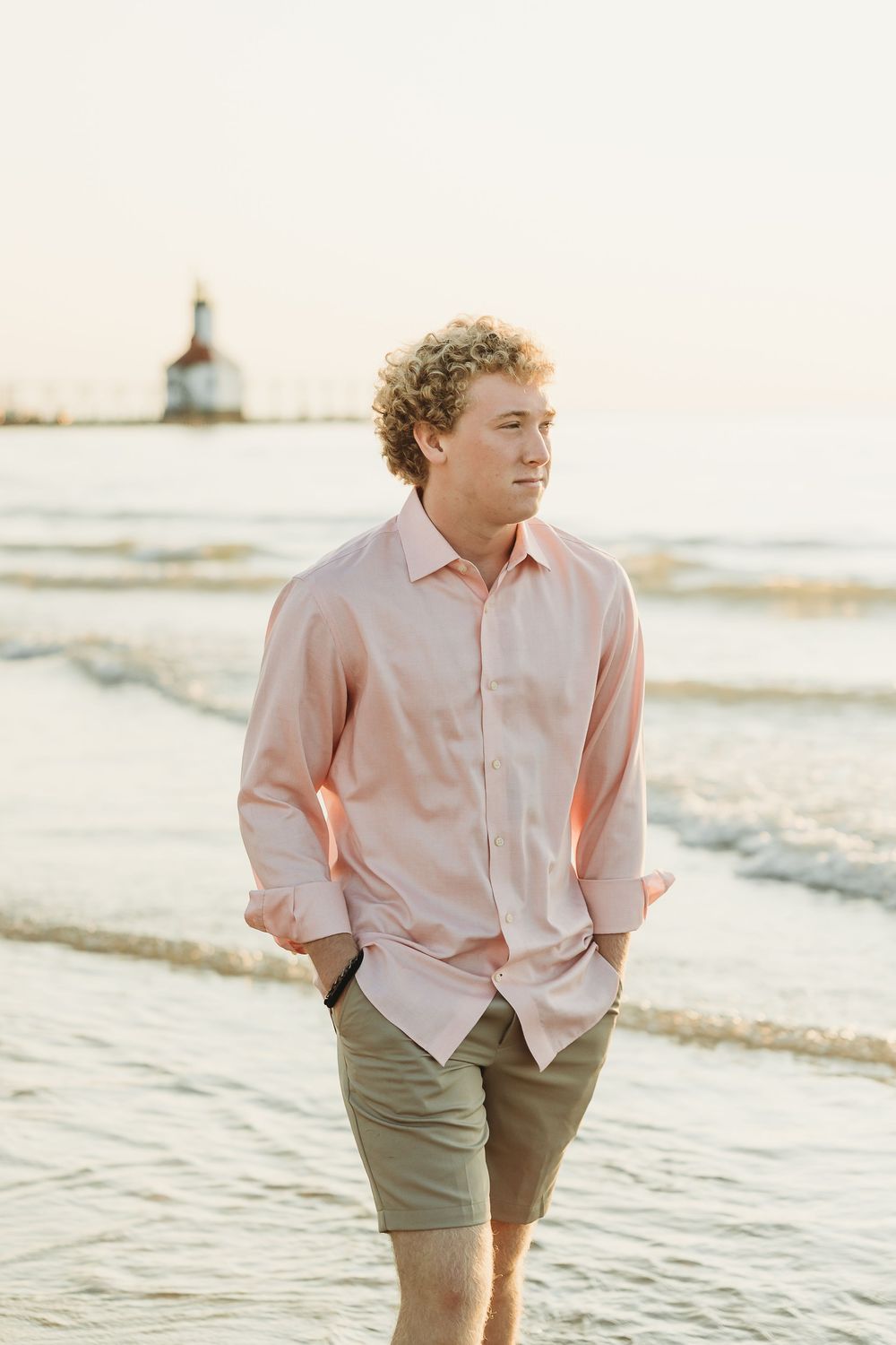 Young man with curly hair in a pink button-up shirt and khaki shorts walks along a sandy beach with a lighthouse in the distance.
