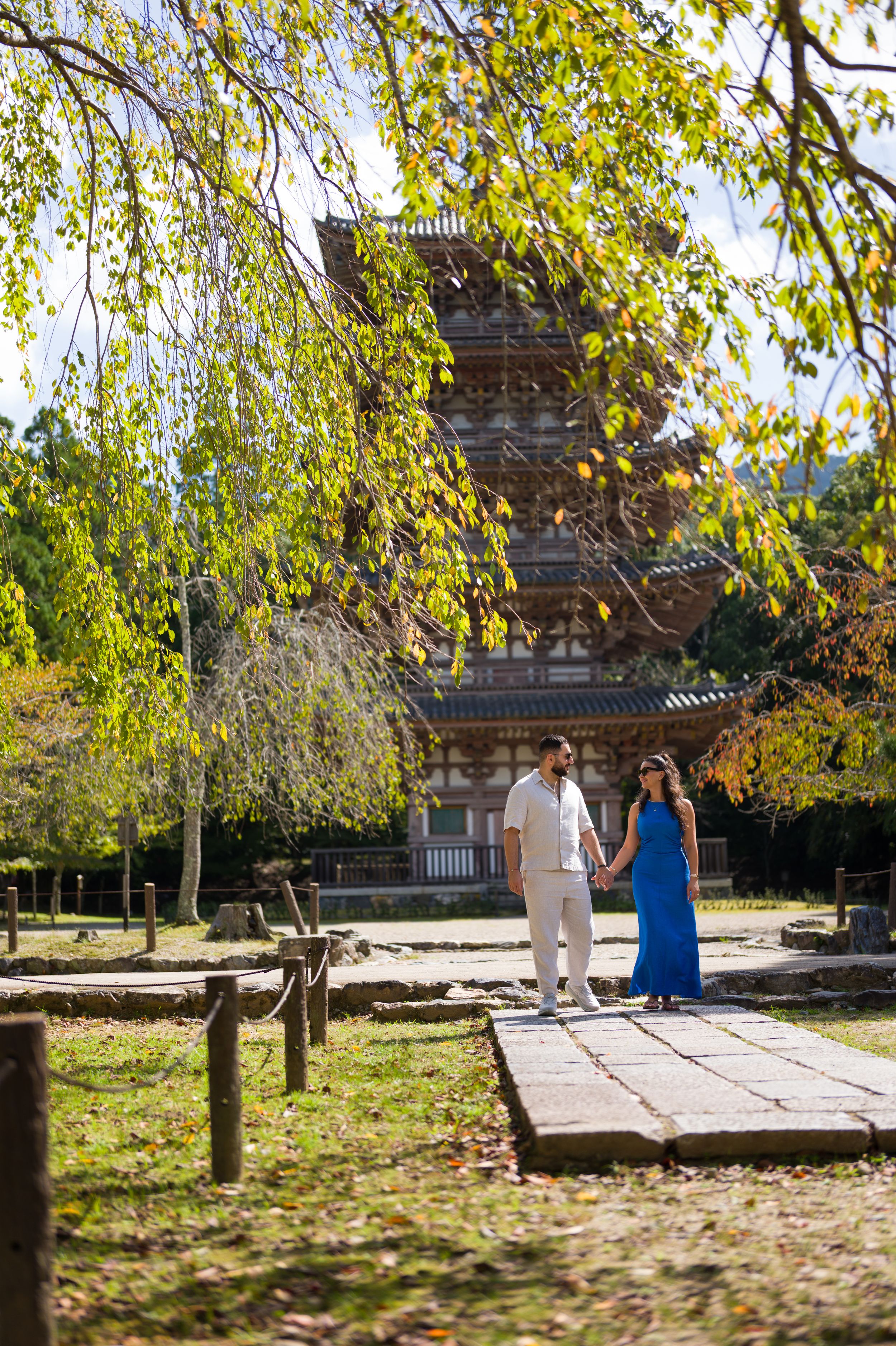 Couple holding hands on a stone path beneath autumn leaves with a historic multi-tiered Japanese wooden pagoda in the background.
