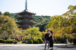 Couple embracing in a sunlit park with autumn foliage in front of a historic multi-tiered Japanese pagoda against a clear blue sky.