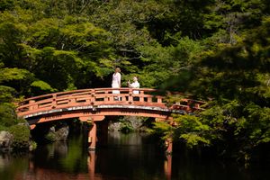 Two people in white stand on a red wooden arched bridge over a calm pond, surrounded by lush green trees in a peaceful Japanese garden.