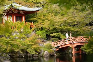 Two people stand on a red wooden arched bridge over a calm pond beside a Japanese temple pavilion surrounded by lush green autumn foliage.