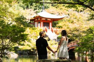 Couple holding hands by a tranquil pond, gazing at a red Japanese pagoda temple surrounded by lush green trees.