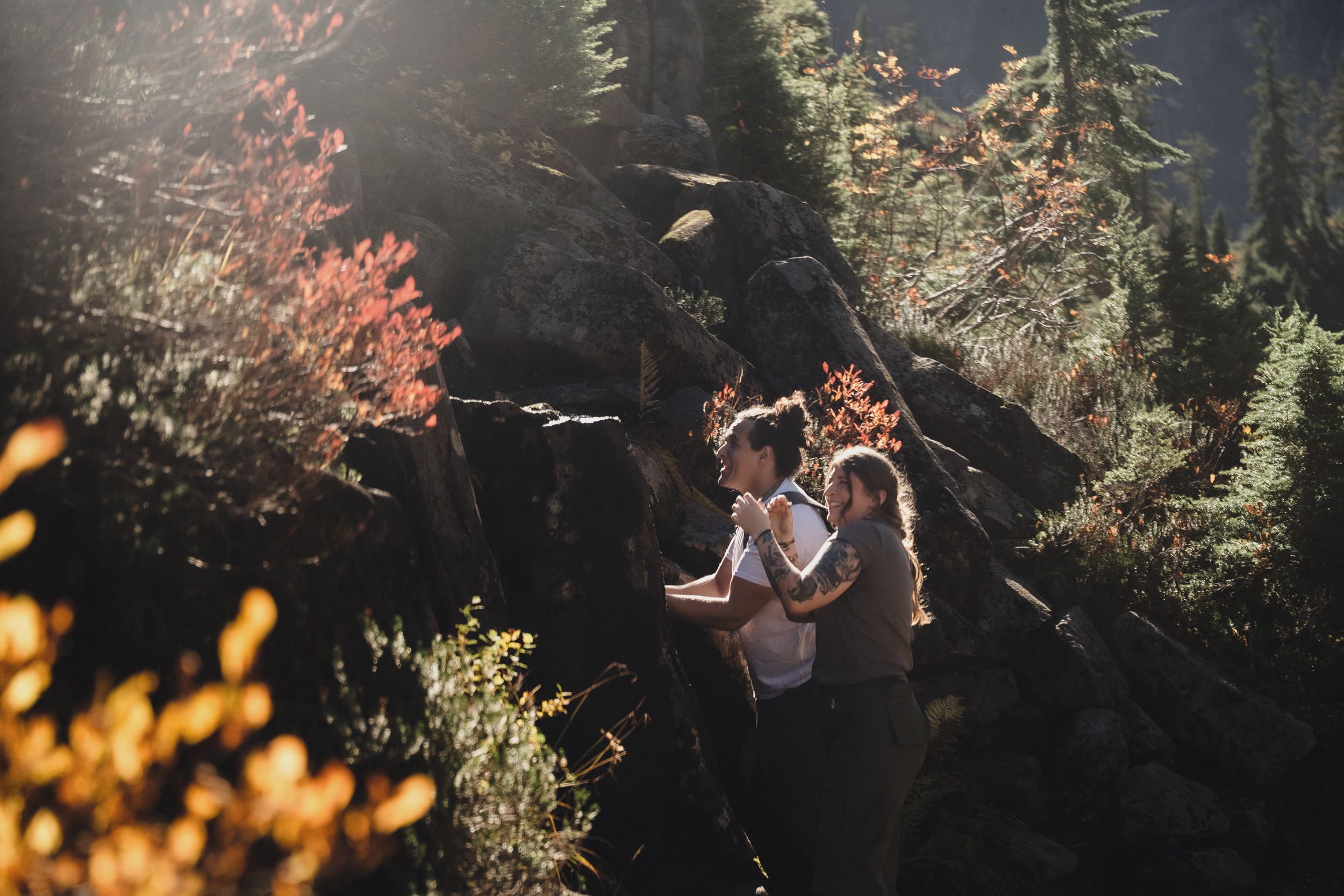 Couple embracing on a rocky mountain trail surrounded by colorful autumn foliage and evergreen trees in warm, golden sunlight.