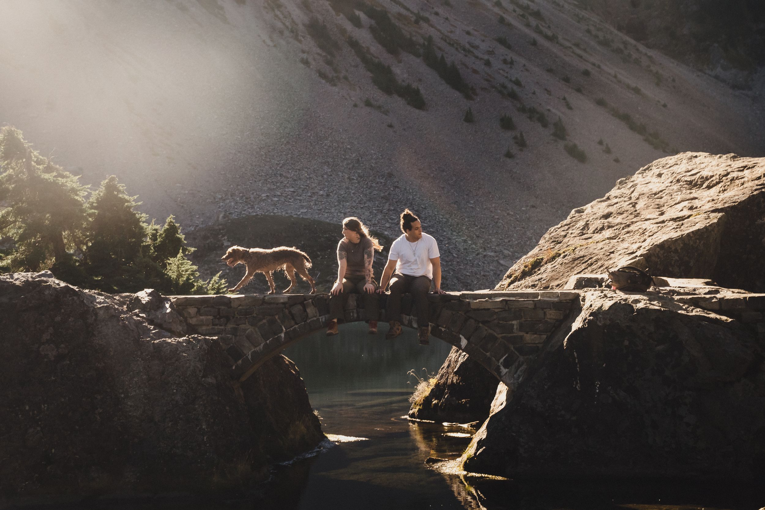 Two people sit on a stone arch bridge over a calm stream while a brown dog stands nearby, with sunlight glowing over rocky mountain terrain.