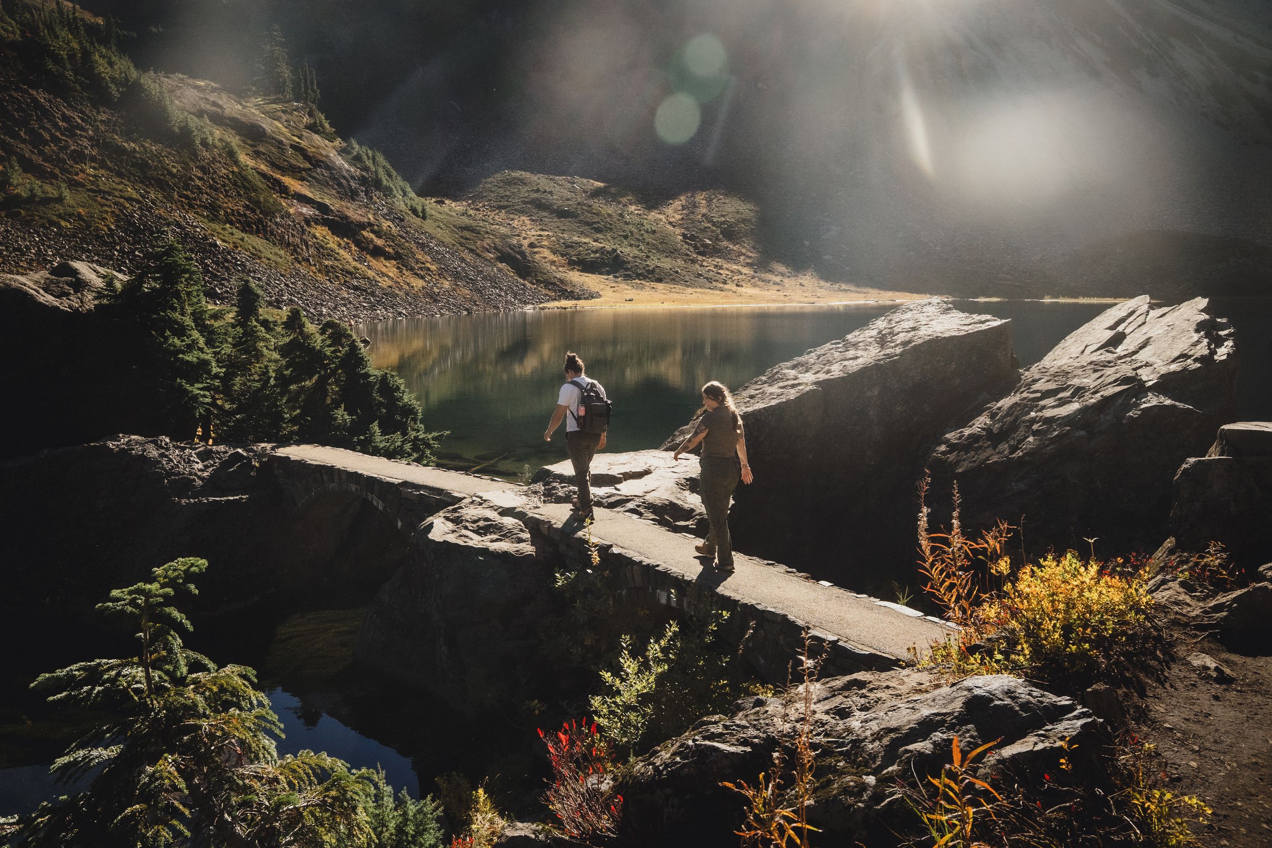 Two hikers walk along a rocky mountain ridge beside a sunlit alpine lake, surrounded by autumn shrubs and rugged cliffs.