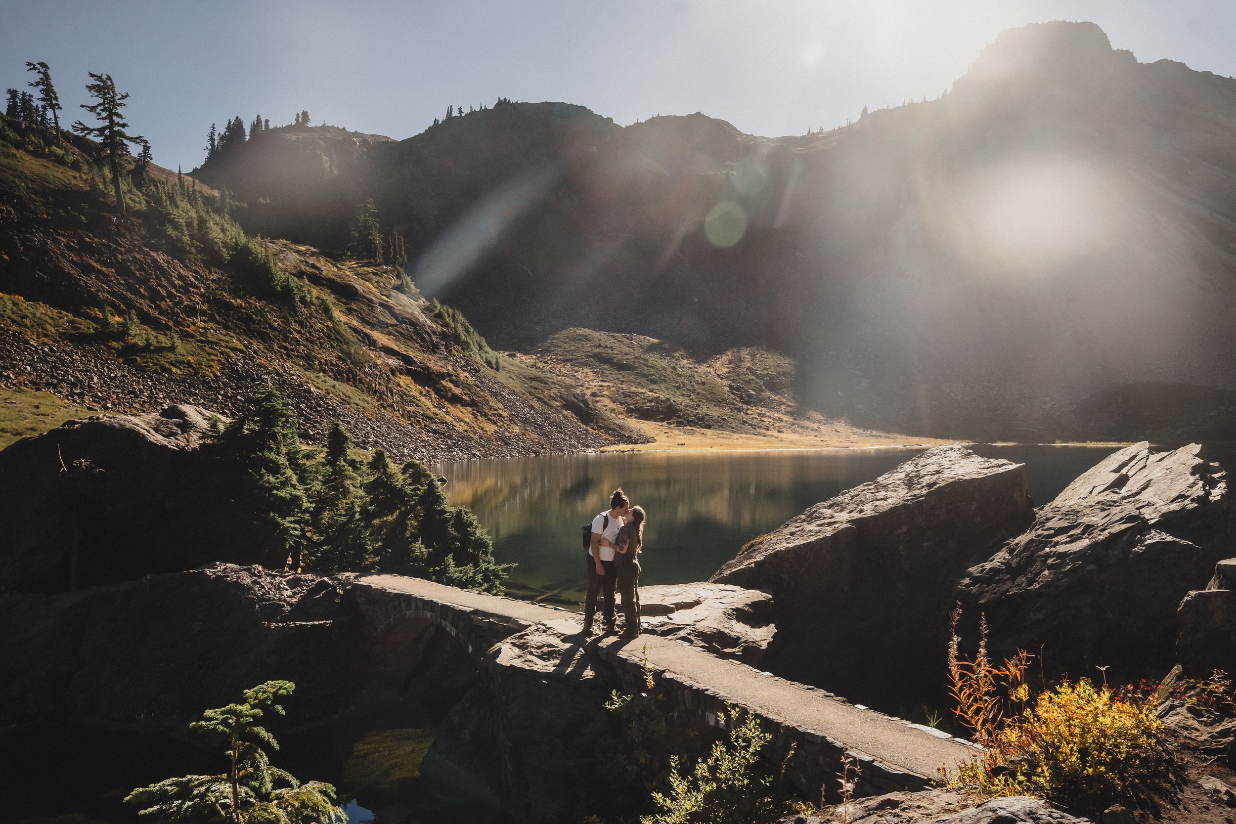 Couple standing together on a rocky ledge beside a calm alpine lake, with sunlit mountains and golden sun flare in the background.
