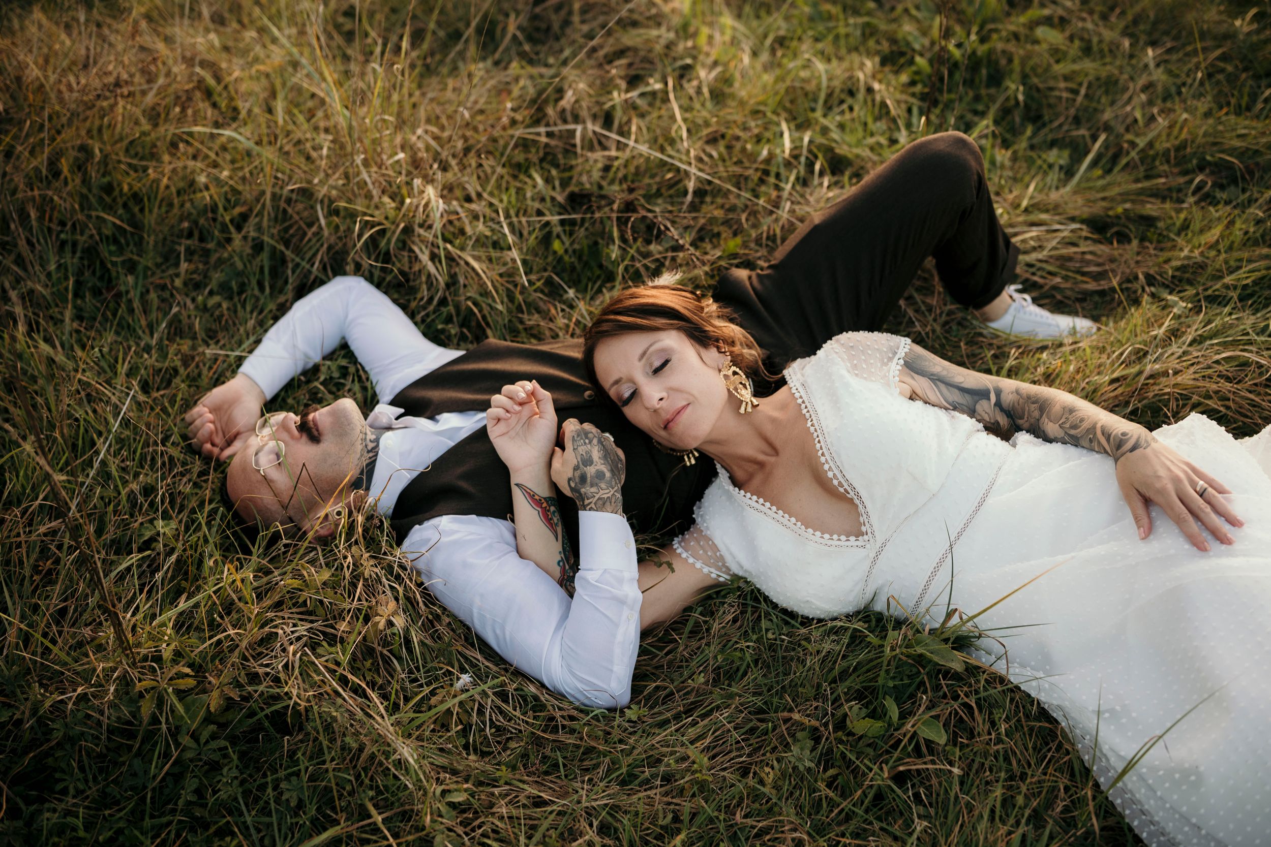 Two people lay in tall grass wearing formal attire during a photo session.
