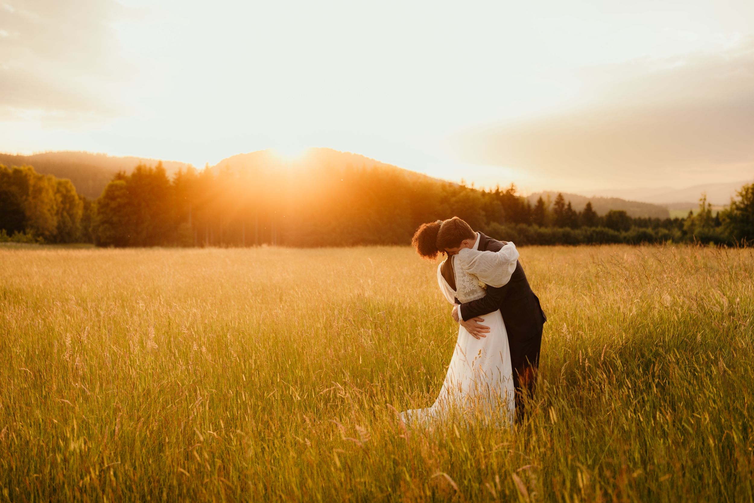 A couple embraces in a golden sunlit meadow with mountains in the background.