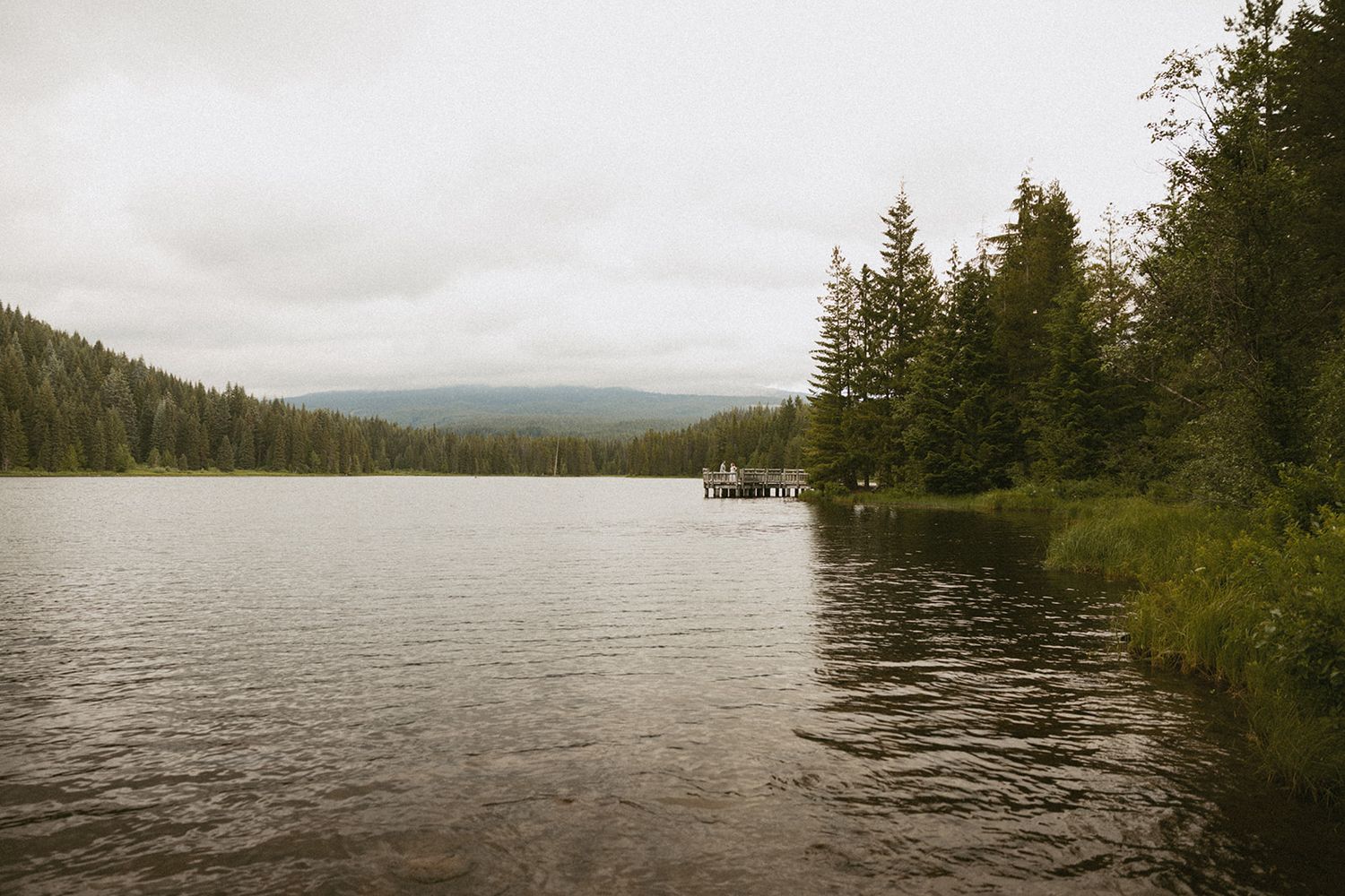 A serene lake surrounded by tall evergreen trees under overcast skies with a small dock visible in the distance.