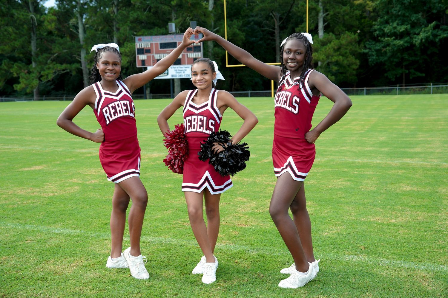 Three cheerleaders in red Rebels uniforms pose on a football field, forming a heart shape with their arms.