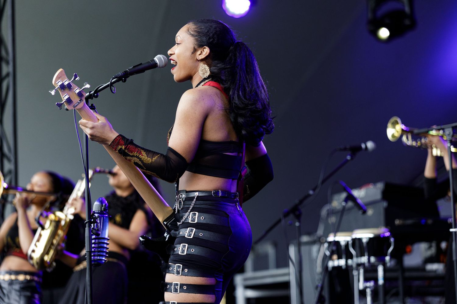 Female singer performing on outdoor stage, holding guitar, wearing black crop top and skirt, with band members behind her.
