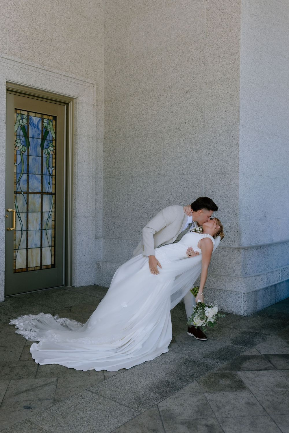 Groom dips bride in a romantic kiss outside a stone building with stained glass door on their wedding day.
