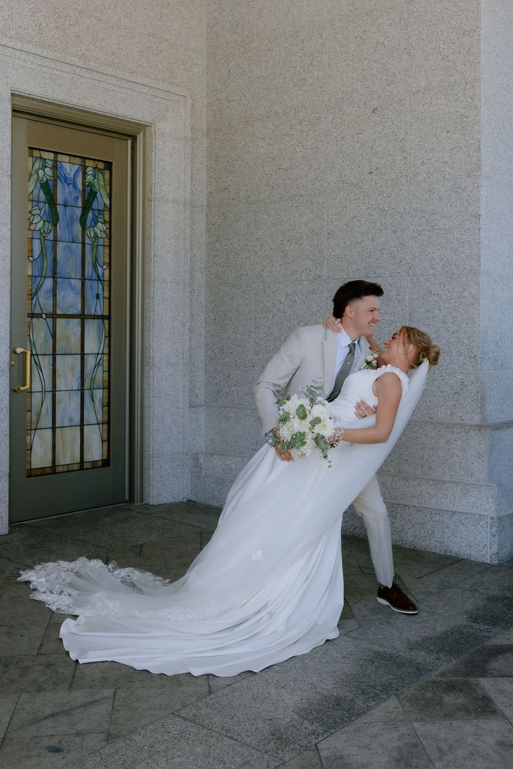 Groom dips bride in white gown holding bouquet outside elegant stone building with stained glass door.