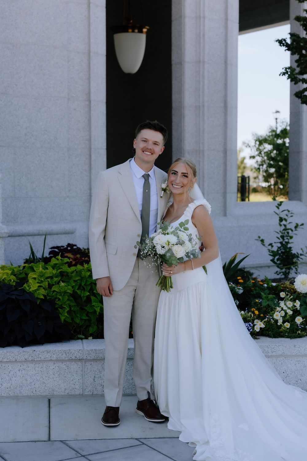 Smiling bride and groom pose outside temple, bride holds white floral bouquet in flowing white gown and beige suit.