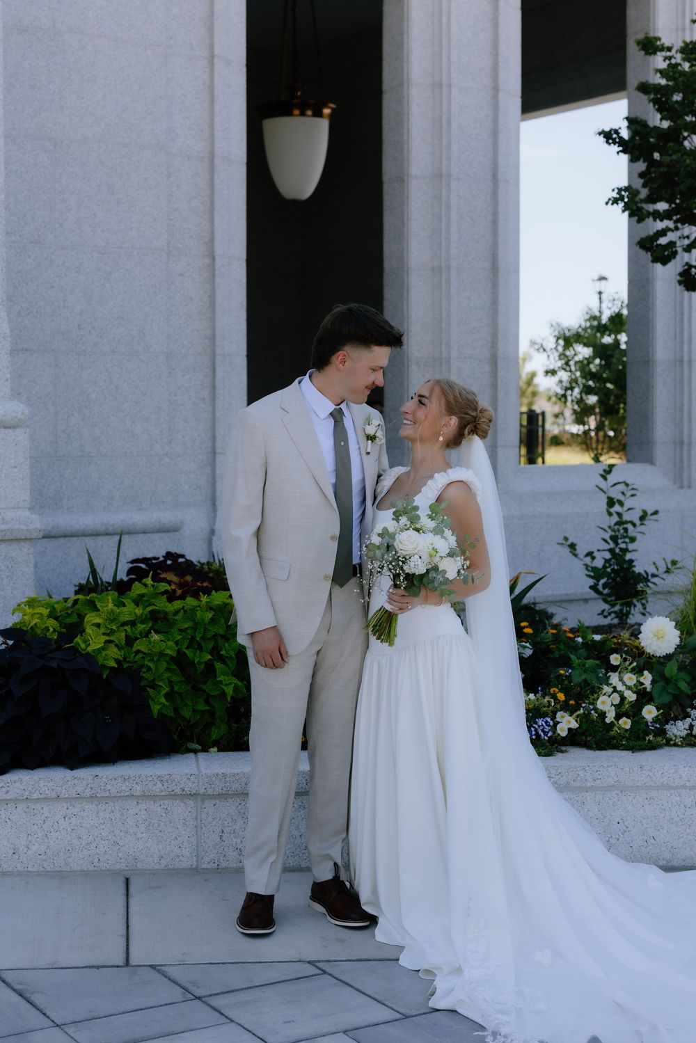 Bride and groom share a loving gaze outside temple columns, bride in white gown holds bouquet, groom in beige suit.