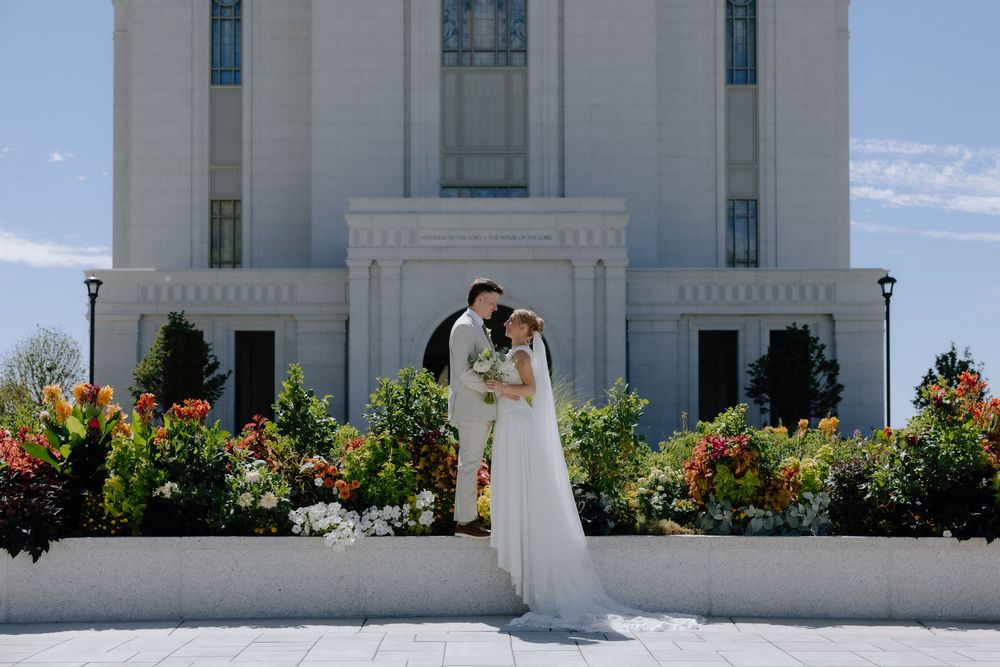 Bride and groom kissing in front of a white temple surrounded by colorful flower gardens on a sunny wedding day.