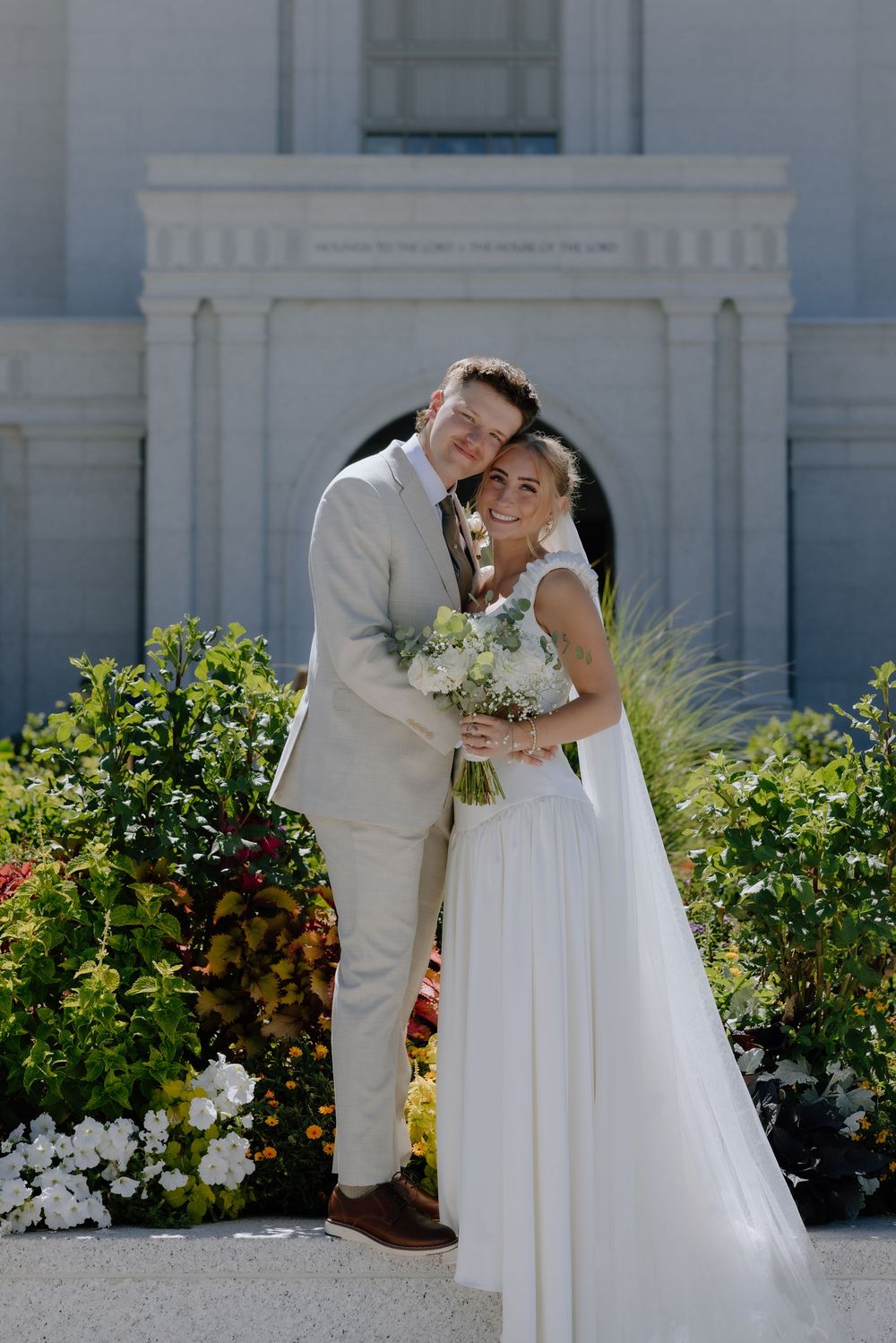 Bride and groom posing in front of a temple surrounded by colorful flowers on their wedding day.