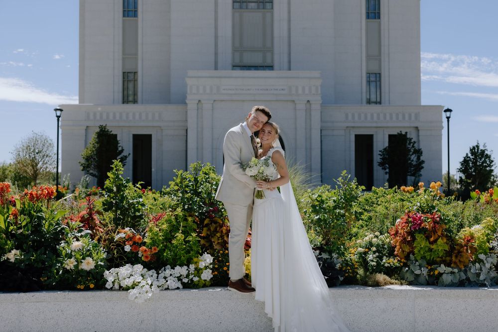 Bride and groom embrace in front of a white temple surrounded by colorful blooming flowers on a sunny wedding day.