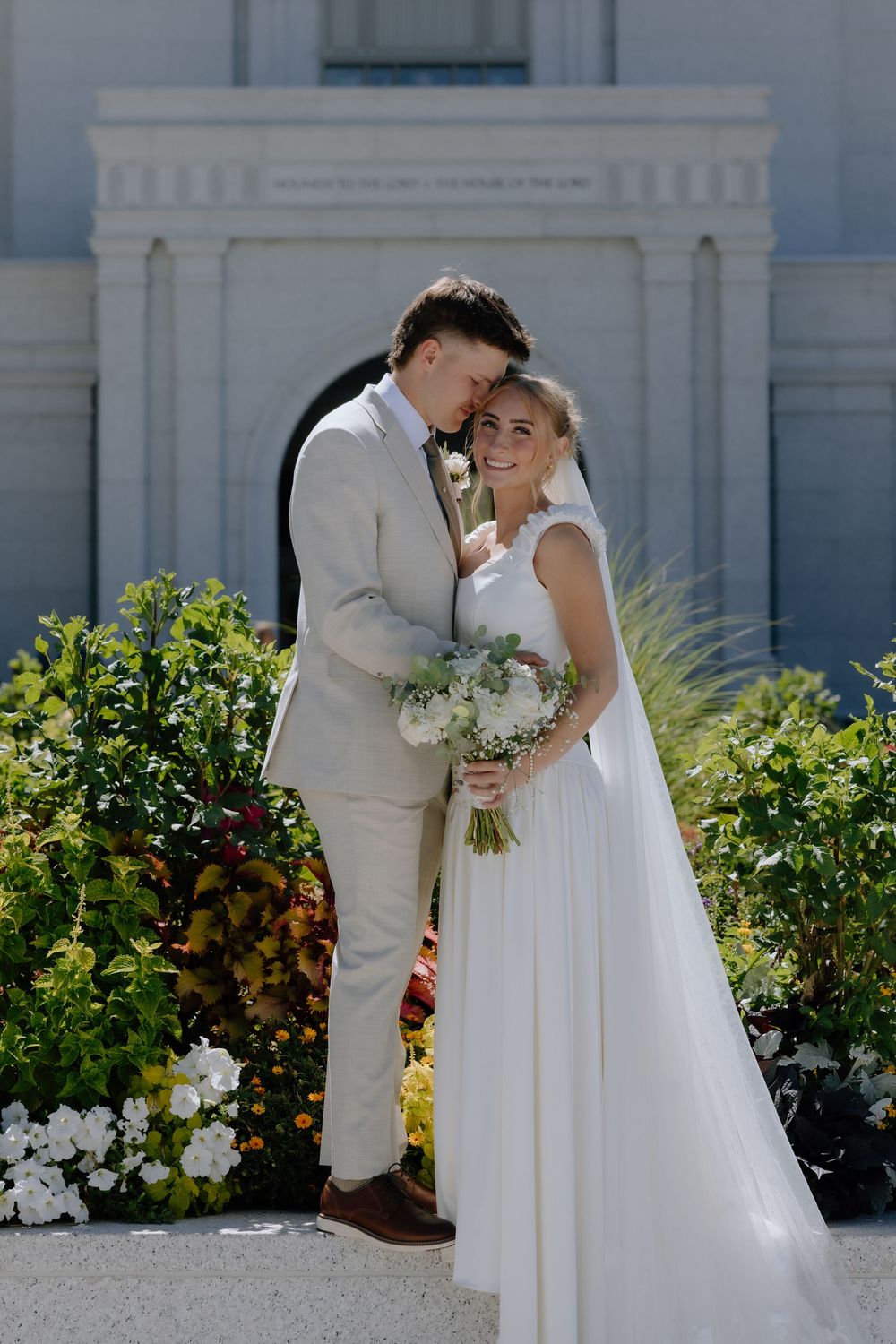 Bride and groom pose together in front of a white building surrounded by lush gardens on their wedding day.