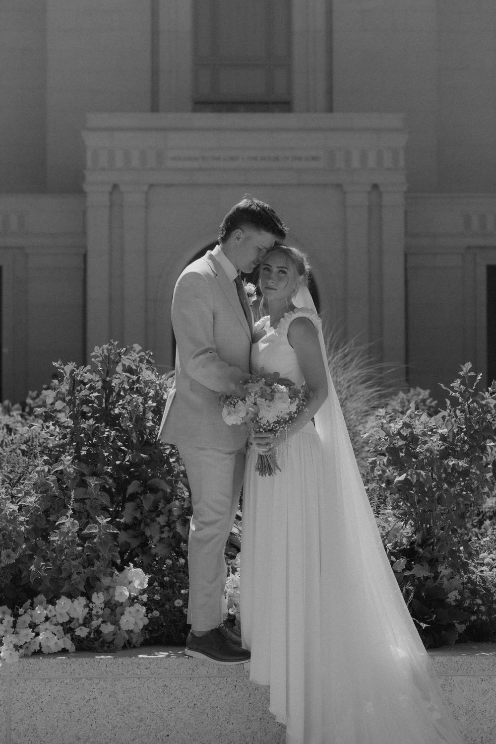 Black and white photo of a bride and groom embracing outdoors near blooming flowers in front of a classic building.