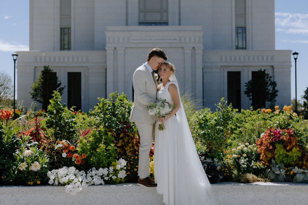 Bride and groom embrace before a white temple surrounded by colorful blooming flowers on their wedding day.