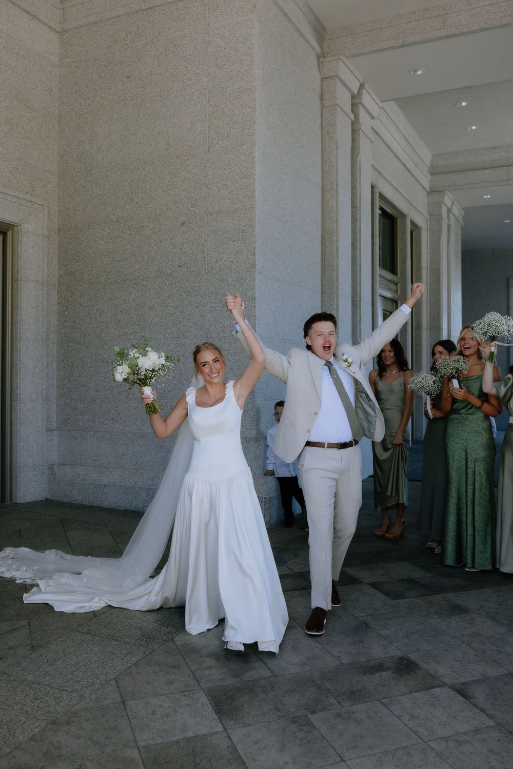 Joyful newlyweds exit wedding venue, bride in white gown holding bouquet, groom in tan suit, bridesmaids in green behind them.