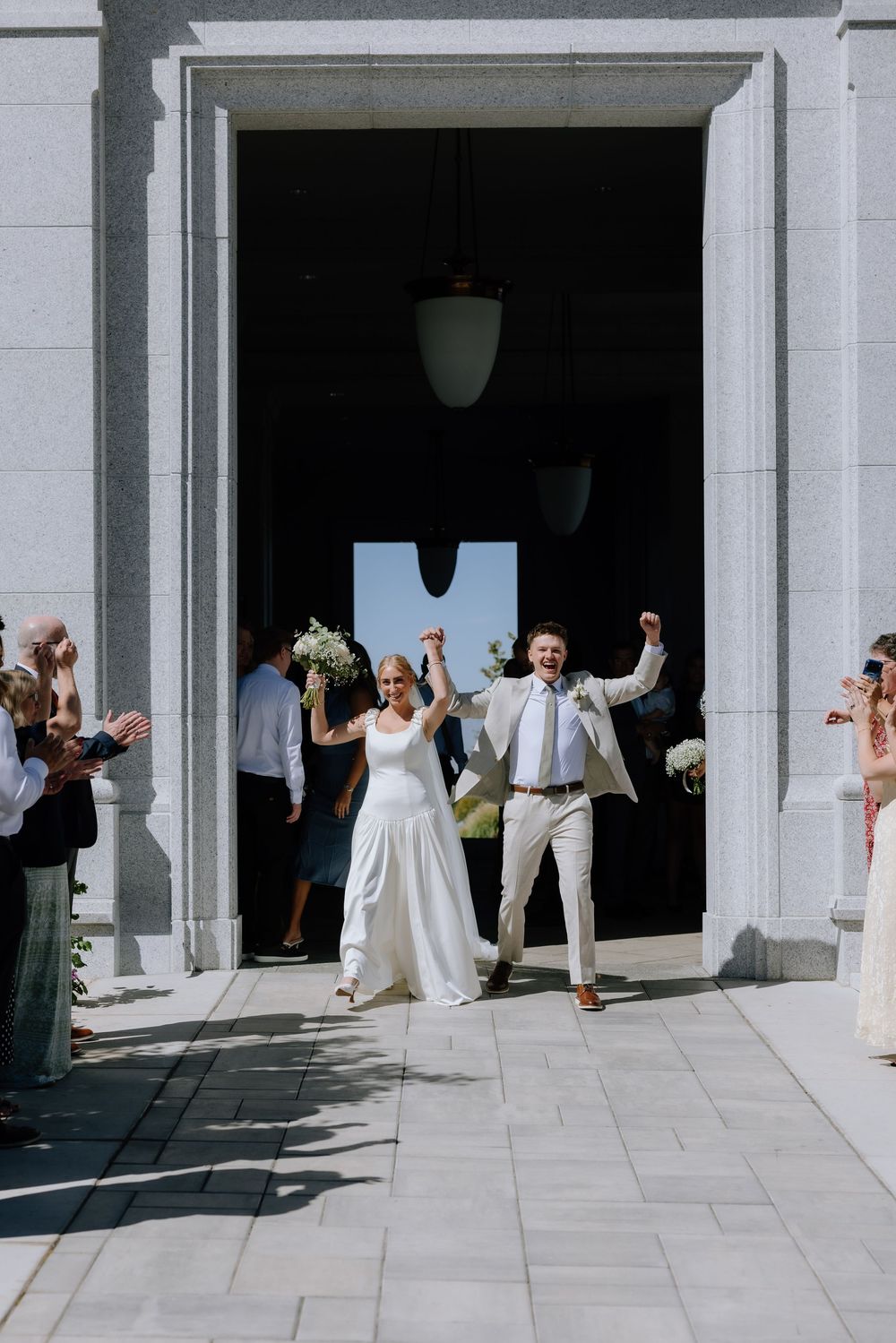 Joyful newlyweds celebrate exiting church doors, bride in white gown holding bouquet, groom in tan suit, guests applauding.