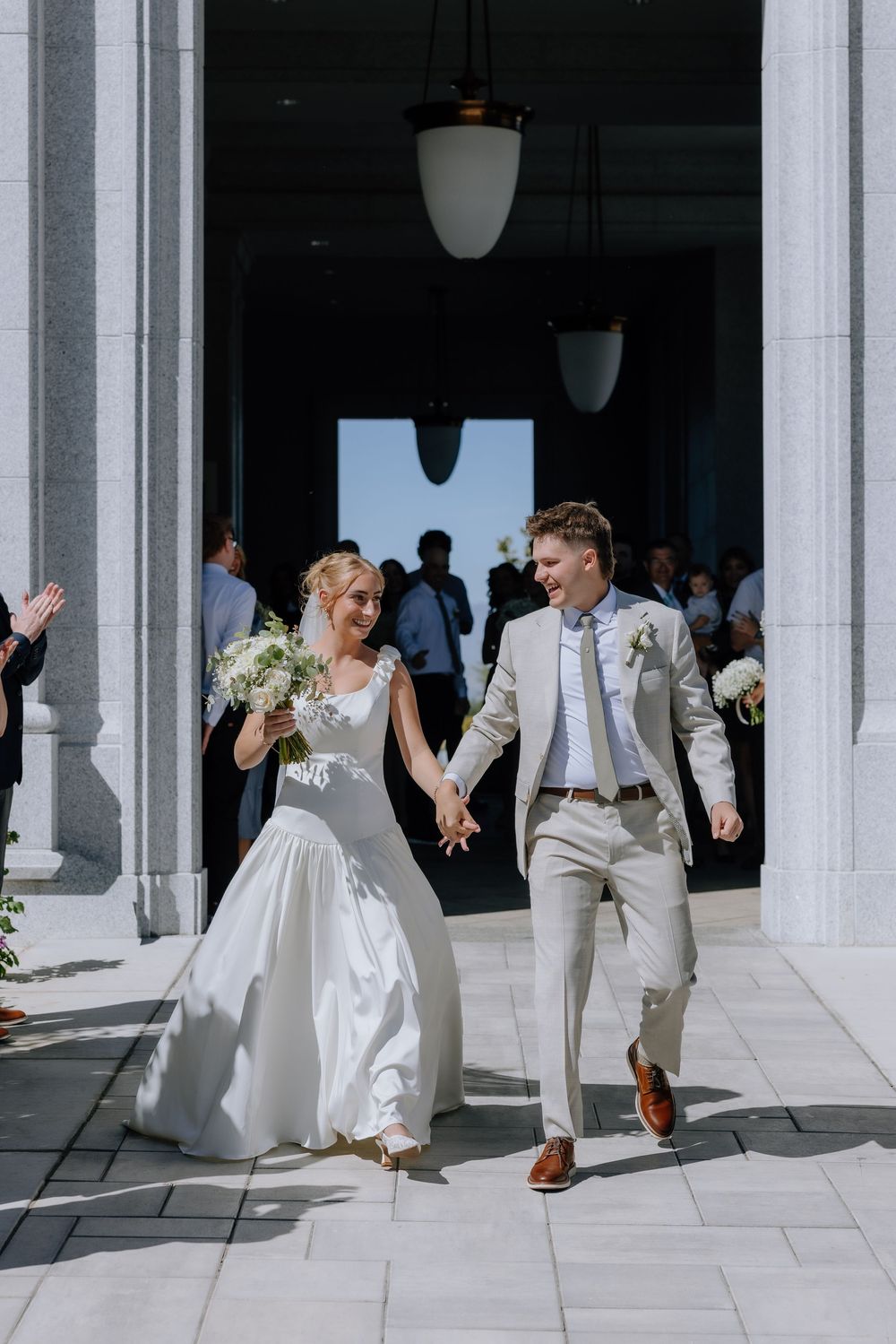 Bride and groom exit church holding hands, bride carries white bouquet in elegant white gown, groom wears beige suit.