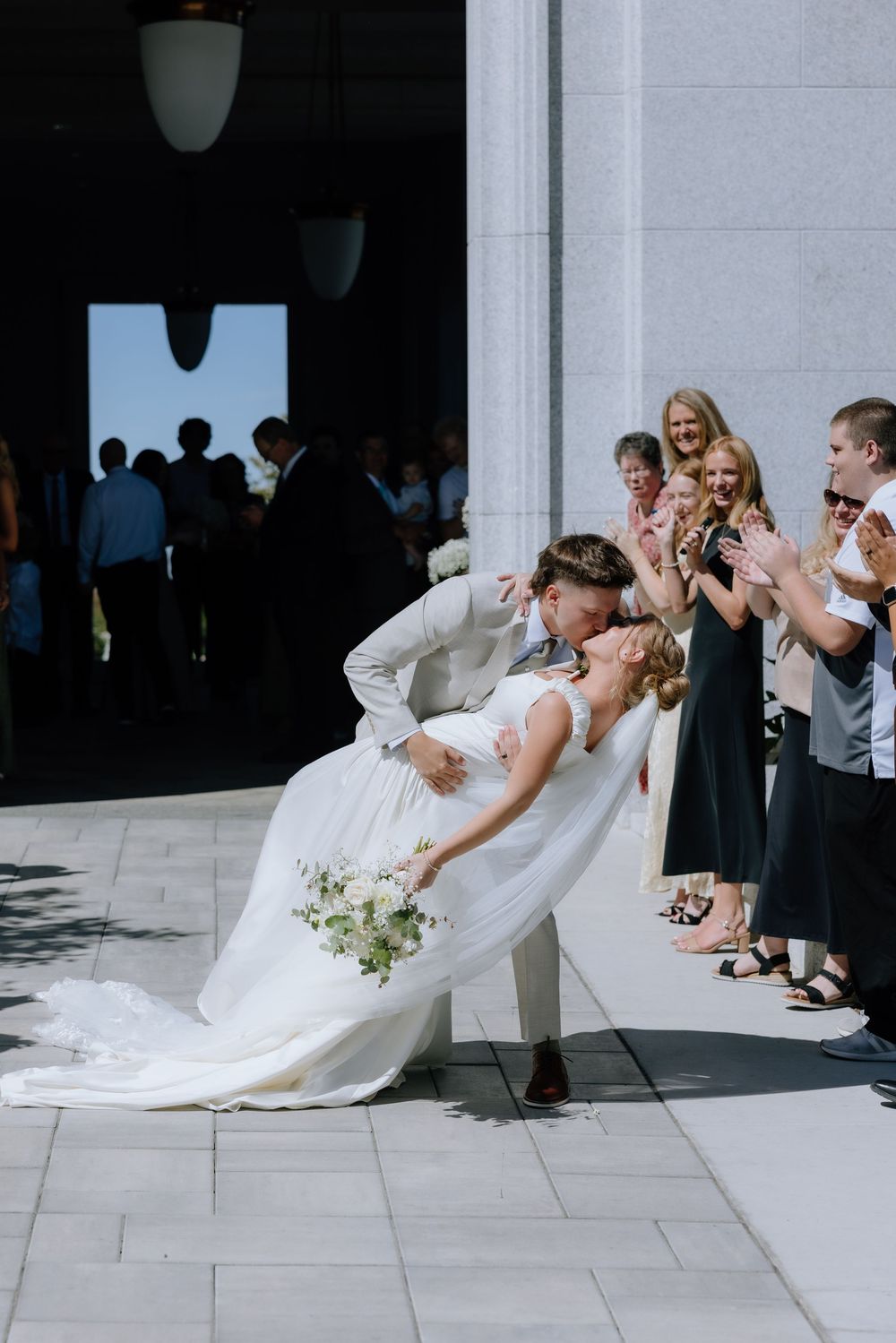 Groom dips bride for a kiss outside wedding venue as guests cheer and applaud in sunny outdoor ceremony.