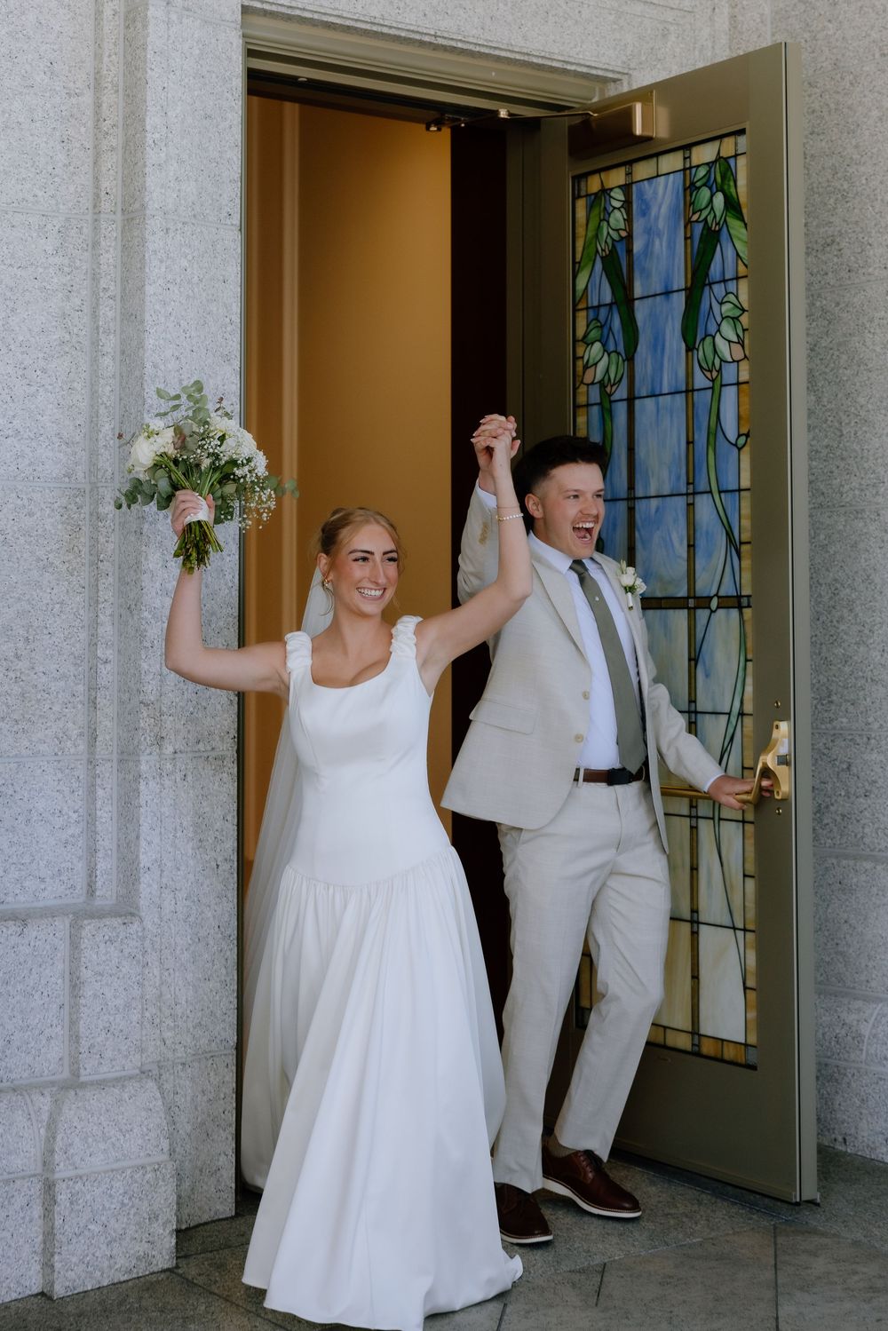 Joyful newlyweds exit church doors, bride in white gown holding bouquet, groom in tan suit, hands raised in celebration.