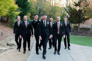 A group of groomsmen in matching black suits and pink ties walk together on a path surrounded by trees.