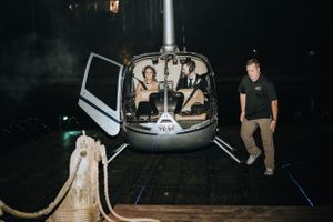 A helicopter sits on a helipad at night with passengers inside and one person standing beside it.