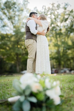 A romantic wedding couple embraces outdoors in a garden setting with blurred white flowers in the foreground.