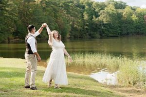 A joyful couple in wedding attire dancing together by a serene lake surrounded by green trees and grass.