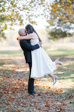 A couple embraces in an autumn setting with fallen leaves, the person in white lifts their foot while being held.