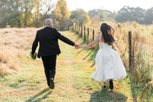 Couple in wedding attire holds hands while walking down rustic path at sunset with golden field and fence.