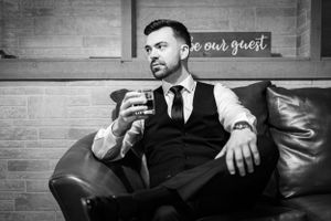 Monochromatic portrait of suited man relaxing on leather couch with drink in hand against brick wall.