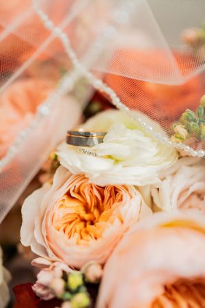 Wedding rings nestled on delicate peach garden roses with flowing white tulle veil in soft lighting.