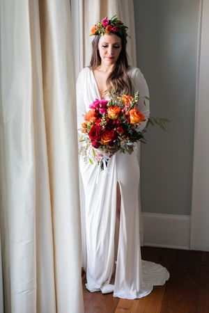 A person in a flowing white wedding dress holds a vibrant autumn bouquet while wearing a floral crown indoors.
