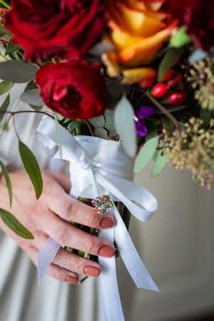 A close-up view of hands holding a bridal bouquet with red roses and a white satin ribbon bow.