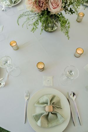 Elegant table setting with sage green napkin, roses in vase, wine glasses and votive candles on white tablecloth.