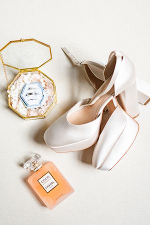 Wedding accessories including white bridal heels, a gold geometric ring box, and peach perfume bottle arranged on white surface.