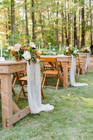 Rustic outdoor wedding ceremony setup with wooden benches decorated with white fabric and floral arrangements in a forest setting.