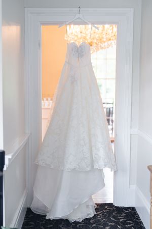 An elegant white lace wedding dress with a sweetheart neckline hangs in a doorway against natural window light.